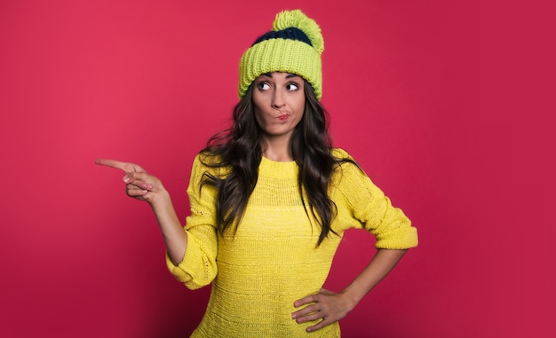 Fascinating woman in a bright yellow sweater and a green hat with pompom is posing in front with her arms folded, looking to the left with a skeptical facial expression