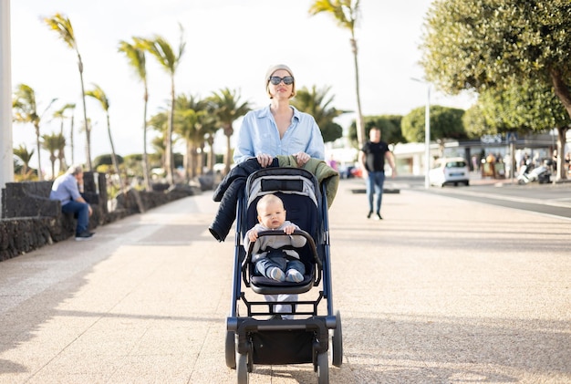 Fashinable mother walking and pushing her infant boy child in baby stroller along palm lined beach promenade of Puerto del Carmen Lanzarote Canary islands Spain