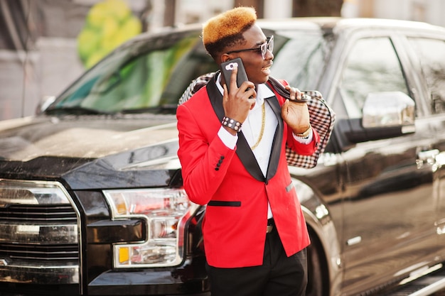 Модная афро-американская модель в красном костюме с яркими волосами позирует на фоне большого черного автомобиля внедорожника и разговаривает по мобильному телефону