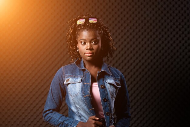 Fashion African Teenager Woman Tan skin black Afro hair beautiful high fashion make up sunglasses pink vast Jean jacket. Egg Crate Studio low lighting shadow Sound Proof Absorbing wall room