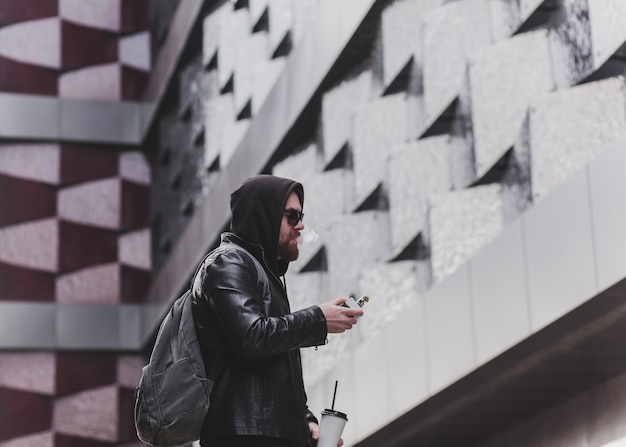 Fashion bearded male dressed in leather jacket, sunglasses and hood vaping. man in holding a mod. A cloud of vapor.