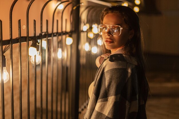 Fashion beautiful hipster girl with glasses and earmuffs in a fashionable plaid shirt walks on the street with light bulbs at night