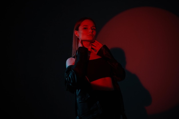 Модная красивая женщина-модель в стильной черной одежде с кожаной курткой на темном фоне в студии с красным творческим светом