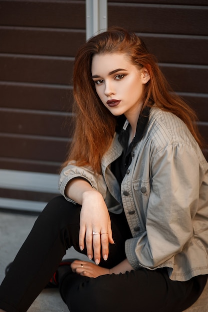 Fashion beautiful young woman in a fashionable jacket and black polo shirt sitting near the gate on the street