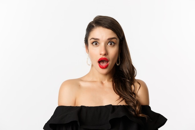 Fashion and beauty concept. Close-up of attractive brunette woman in black dress open mouth surprised, looking in awe at camera, white background