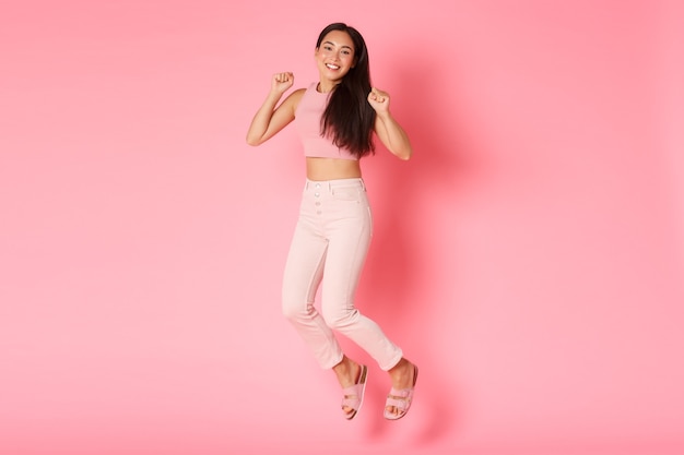 Fashion, beauty and lifestyle concept. Carefree beautiful asian girl jumping from happiness, looking upbeat and relaxed, celebrating vacation, enjoying shopping, standing pink wall.