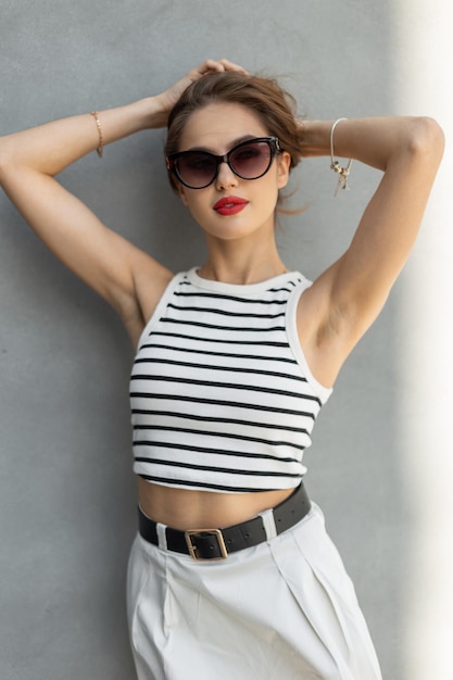 Photo fashion beauty pretty woman with red lips with fashionable sunglasses in stylish summer casual outfit stands and poses near a gray wall on the street