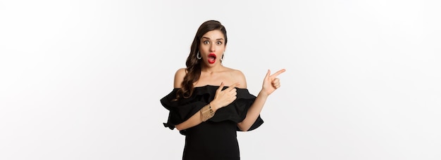 Fashion and beauty surprised woman in black glamour dress pointing fingers left showing advertisemen