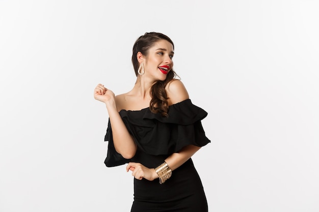 Fashion and beauty. Woman celebrating and dancing in black dress, having fun and smiling, standing over white background