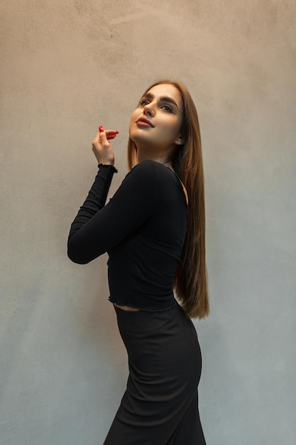 Модная красавица-модель в черном элегантном наряде с топом и брюками стоит возле серой стены в помещении