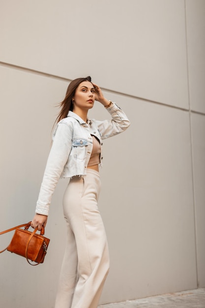 Fashion beauty young woman in trendy jeans clothes with stylish leather handbag walks in the city near gray building