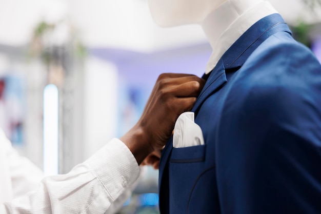 Fashion boutique african american worker hands adjusting formal jacket on model, displaying clothes for sale. Shopping mall assistant fastening male costume on mannequin close up