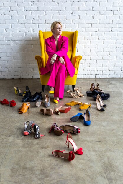 Fashion business lady in bright pink outfit sitting yellow armchair at loft room random many shoes