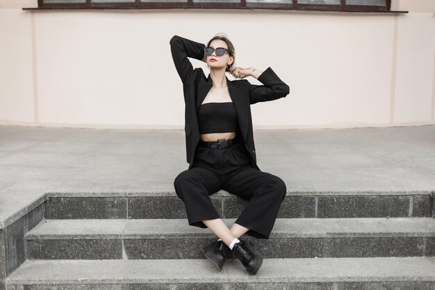 Fashion business pretty girl with sunglasses in a fashionable black elegant suit outfit with jacket top pants and shoes sits on the steps outside the building