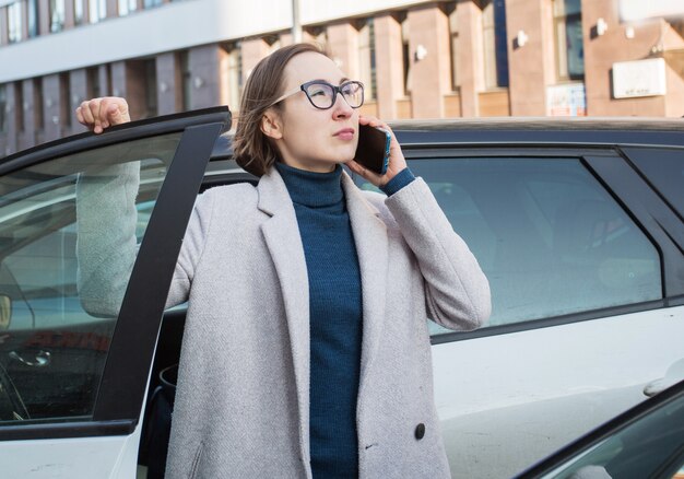 Модная бизнес-леди в очках с телефоном выходит из машины.