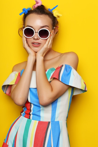 Мода прохладная женщина позирует в солнечных очках на желтом. Битник молодая женщина с бигуди в волосах