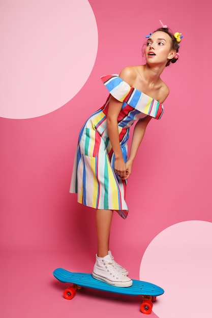 Fashion cool woman posing with a skateboard, multicolour dress and sneakers on pink.