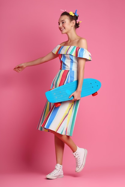 Fashion cool woman posing with a skateboard, multicolour dress and sneakers on pink.