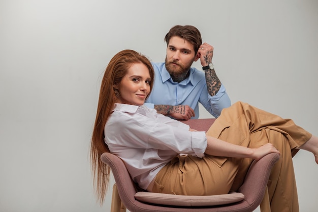 Fashion couple of lovers Beautiful happy redhead girl model with red hair sits and rest in a chair and handsome brutal hipster man with beard and tattoo