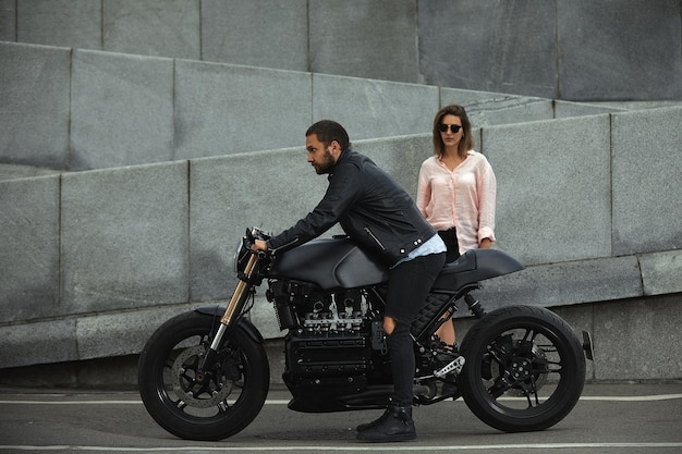 バイクに座っているファッションカップル、背景の石の壁。現代のオートバイを持つ若い男と女。