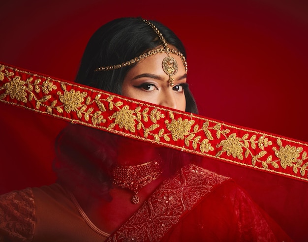 Fashion culture and portrait of Indian woman with veil in traditional clothes jewellery and sari Religion beauty and eyes of female person on red background with accessory cosmetics and makeup