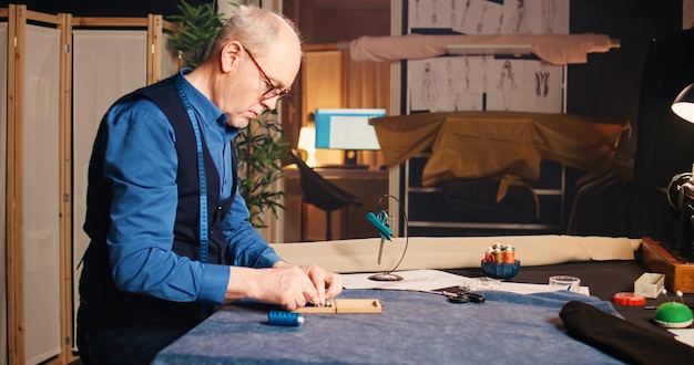 Fashion designer choosing items and tools to create custom made clothes, he has measuring tape around his neck. Old tailor crafting handmade clothing design in workshop atelier. Handheld shot.