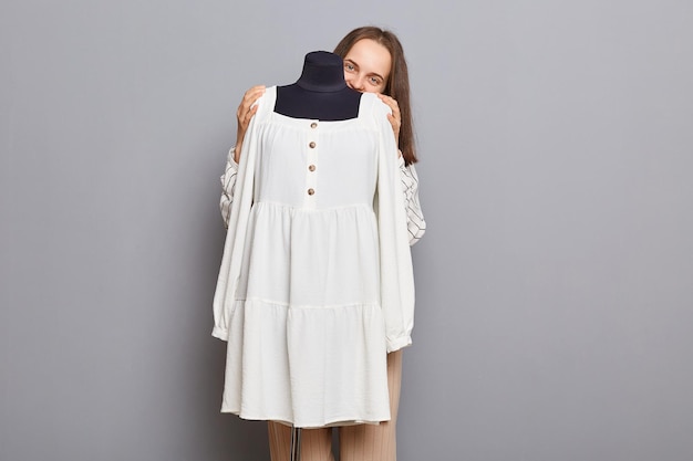 Fashion designer entrepreneur Creative clothing workshop Cute smiling positive woman sewer standing hiding behind new dress on mannequin iisolated over gray background