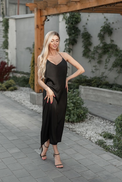 Fashion elegance pretty woman blonde model with smile in black strappy long dress with heels shoes walks on the street