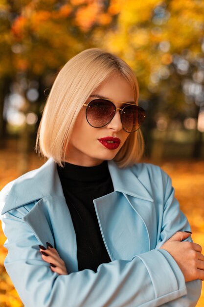 Fashion fall portrait of a beautiful business woman with vintage cool sunglasses in a blue coat in nature on a background of colorful autumn leaves