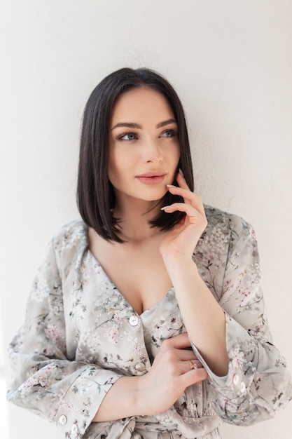 Fashion fresh spring female portrait of beautiful woman with hairstyle in fashionable flowers dress stands and poses near a white wall on the street