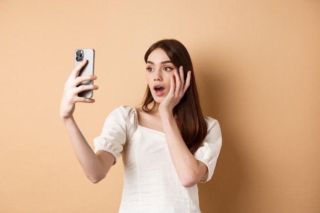 Fashion girl record smartphone blog, taking selfie on cellphone, standing on beige background.