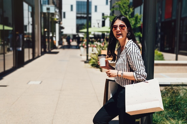 Moda ragazza in occhiali da sole in piedi all'aperto rilassante riposando godetevi la giornata di sole dopo lo shopping nel centro commerciale di stanford. bella giovane signora asiatica che tiene borse caffè sorridente con gioia sotto il sole.