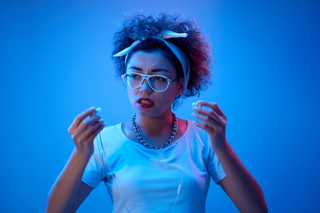 Fashion girl with afro curls and a disgruntled face uses earphones on a neon wall