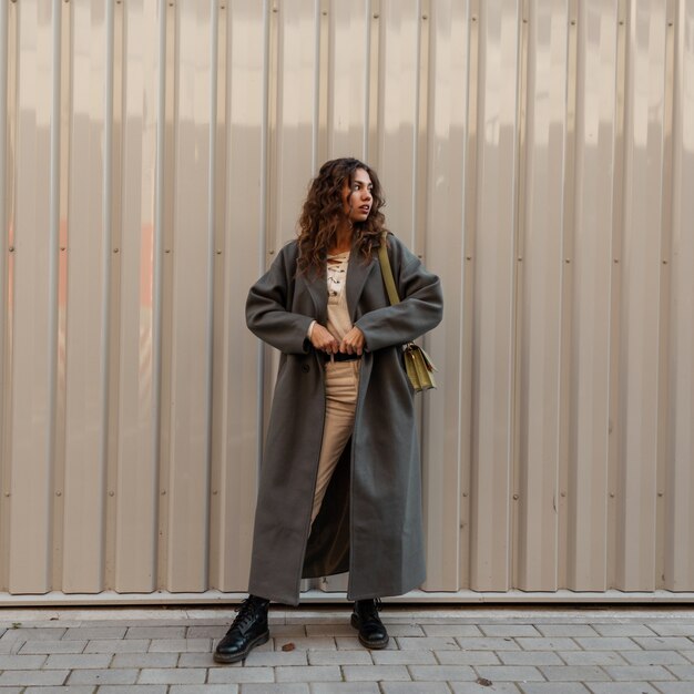 Fashion girl with curly hair in a vintage green long coat with a handbag stands near a metal wall. Urban feminine style and beauty