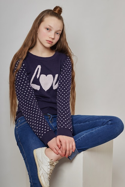 Fashion girl with long hair in casual clothes sitting on a white cube and posing. Beautiful young