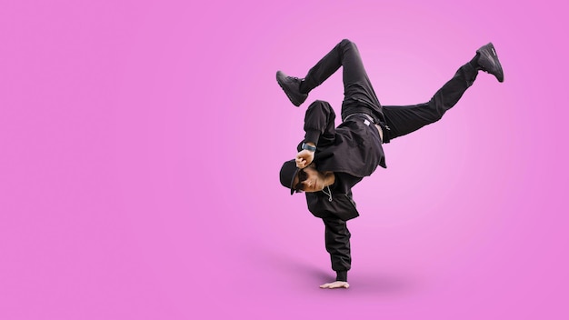Фото Модный красивый танцор в стильной бейсбольной кепке и солнцезащитных очках в модном черном наряде с джинсами-бомберами и кроссовками выступает на розовом фоне и стоит под рукой танцевальный мальчик