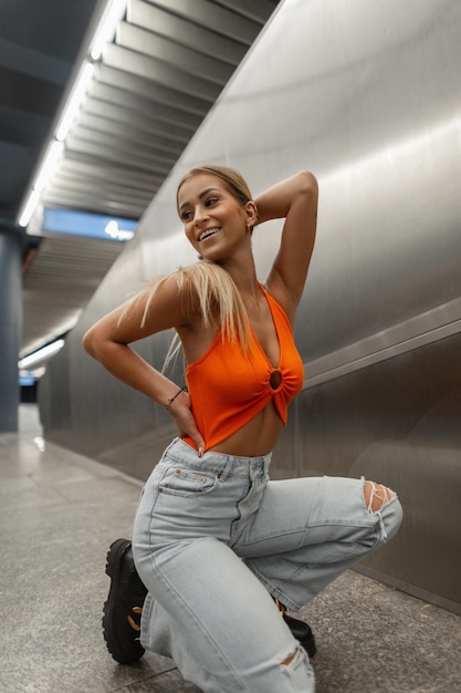오렌지색 섹시한 탑과 빈티지한 파란색 찢어진 청바지에 미소를 짓고 있는 패션 행복한 아름다운 소녀가 금속 도시 배경의 지하철에 앉아서 포즈를 취합니다.
