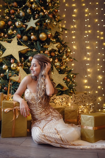 Fashion interior photo of beautiful sensual woman with blond hair in luxurious golden dress posing beside a Christmas tree. Wish you a Merry Christmas.