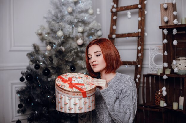 Fashion interior photo of beautiful young woman with red hair and charming smile, wears cozy knitted cardigan, posing beside Christmas tree and presents with chocolate cake with asterisks