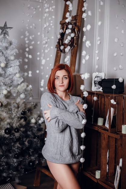 Fashion interior photo of beautiful young woman with red hair and charming smile, wears cozy knitted cardigan, posing beside Christmas tree and presents with chocolate cake with asterisks