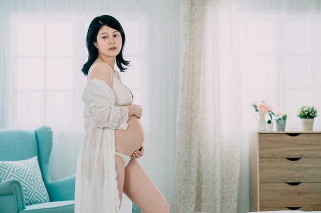 fashion interior of side view beautiful pregnant asian chinese woman with dark hair in elegant lingerie and lace robe standing by armchair in bedroom. young future parenthood girl hold naked belly