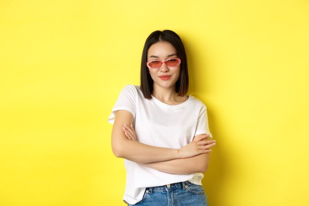 Fashion and lifestyle concept. Sassy and confident asian woman in trendy sunglasses looking self-assured at camera, standing over yellow.