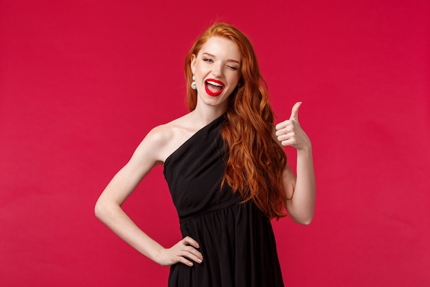 Fashion, luxury and beauty concept. Portrait of sassy gorgeous young redhead woman in black dress, wink at with thumb-up, recommend or approve something really awesome, likes party