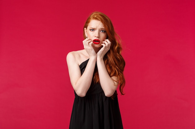 Fashion, luxury and beauty concept. Portrait of timid and scared ginger girl in black dress feel unsafe or insecure, touch face frowning frightened, look concerned and worried , on a red wall