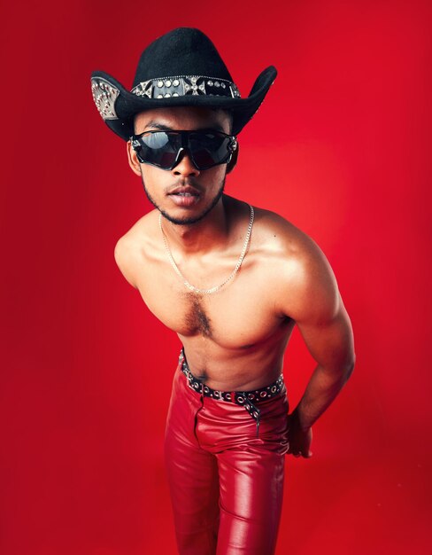 Fashion man and cowboy in hat shirtless with sunglasses and gen z isolated on red background confident model cool and leather pants with eyewear accessory western aesthetic and edgy in studio