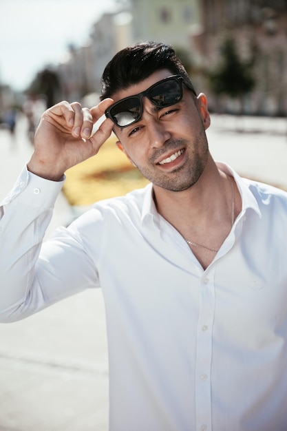 Fashion man in a white shirt is standing on the street at a sunny day is posing watching holding glasses