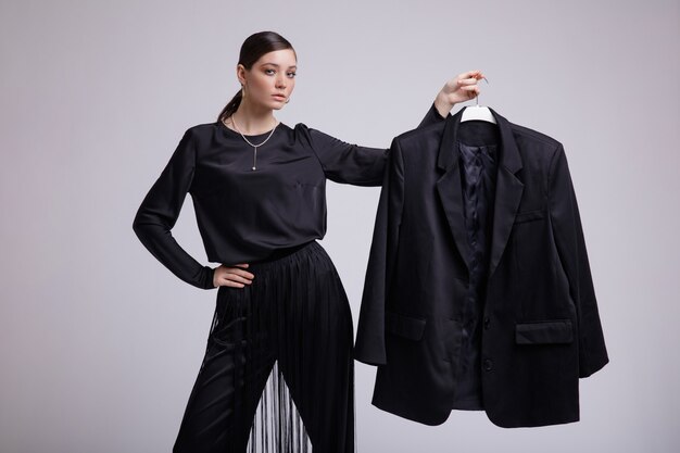 Fashion model in black blouse fringe pants on gray background Holds jacket in hands on a hanger