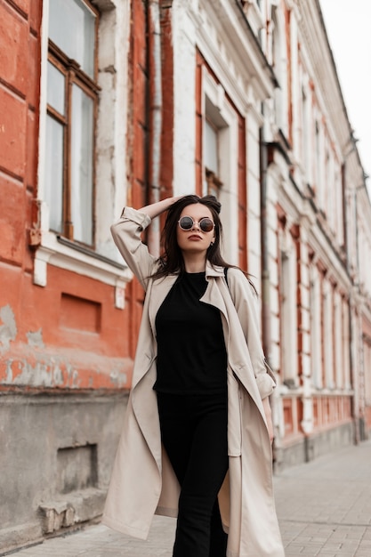 Fashion model comely young woman in fashionable sunglasses in spring vintage trench coat in stylish black wear walks on city near old building. Trendy girl brown-haired travels on street. Casual look.