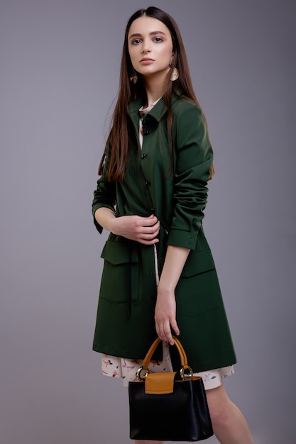 Фотомодель в зеленом пальто с сумкой красивая молодая женщина Студия выстрел Серый фон