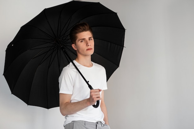 Fashion model handsome young man in youth fashionable t-shirt
stands with vintage umbrella near wall in room. sexy modern nice
guy in summer wear posing with black umbrella on white background
indoors